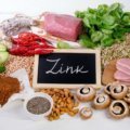 Verschiedene zinkhaltige Lebensmittel wie Fleisch, Meeresfrüchte, Nüsse, Samen, Hülsenfrüchte, Avocado und Spinat liegen auf einem Tisch, daneben ein Schild mit der Aufschrift „Zink“.