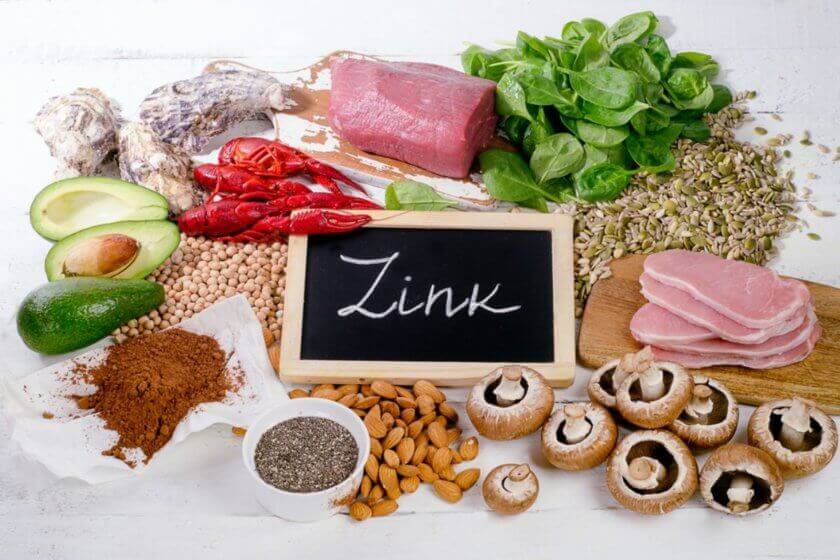 Verschiedene zinkhaltige Lebensmittel wie Fleisch, Meeresfrüchte, Nüsse, Samen, Hülsenfrüchte, Avocado und Spinat liegen auf einem Tisch, daneben ein Schild mit der Aufschrift „Zink“.