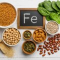 Eisen / Eisenhaltige Lebensmittel wie Linsen, Kichererbsen, Spinat, Kürbiskerne, Sonnenblumenkerne, Mandeln und Quinoa rund um eine kleine Tafel mit dem Symbol ‚Fe‘ auf hellem Holz.