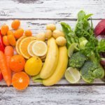 Farbcode der Ernährung & bunt essen (Buntes Arrangement aus Obst und Gemüse in verschiedenen Farben – Tomaten, Karotten, Zitrusfrüchte, Bananen, Brokkoli, Trauben und rote Beete auf hellem Holzhintergrund.)
