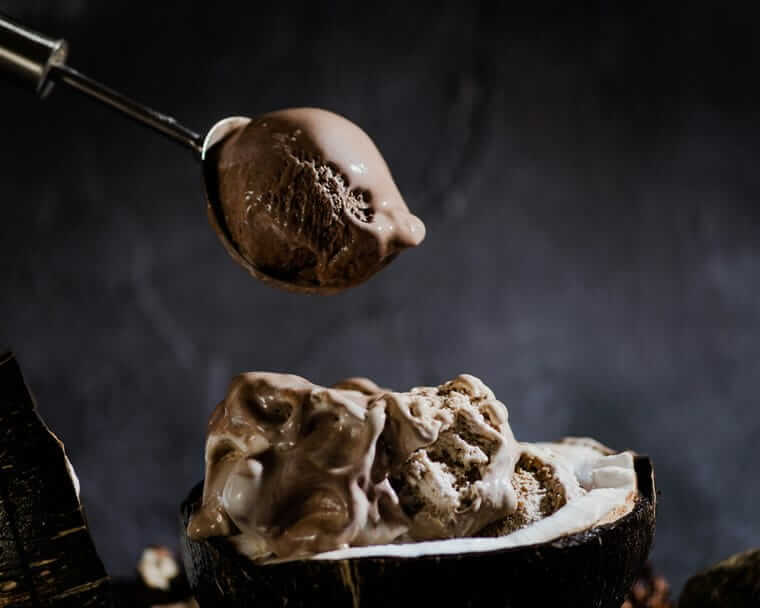 Der kleine Löffel-Besteck-Guide: Welchen brauche ich wofür? Eislöffel / Ein Löffel mit cremigem Schokoladeneis schwebt über einer halbierten Kokosnuss, die mit geschmolzenem Schokoladeneis gefüllt ist, vor einem dunklen Hintergrund.