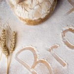 Brotlaib auf bemehlter Oberfläche mit Ähren und dem Wort „Brot“ im Mehl – Symbol für Natürlichkeit, Frische und handwerkliche Qualität beim Brotbacken