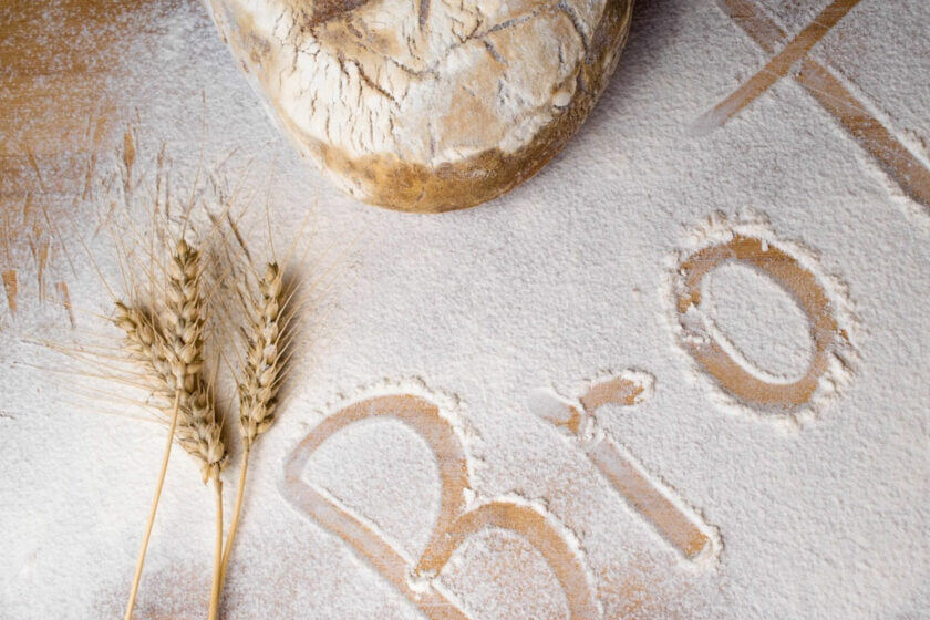 Brotlaib auf bemehlter Oberfläche mit Ähren und dem Wort „Brot“ im Mehl – Symbol für Natürlichkeit, Frische und handwerkliche Qualität beim Brotbacken