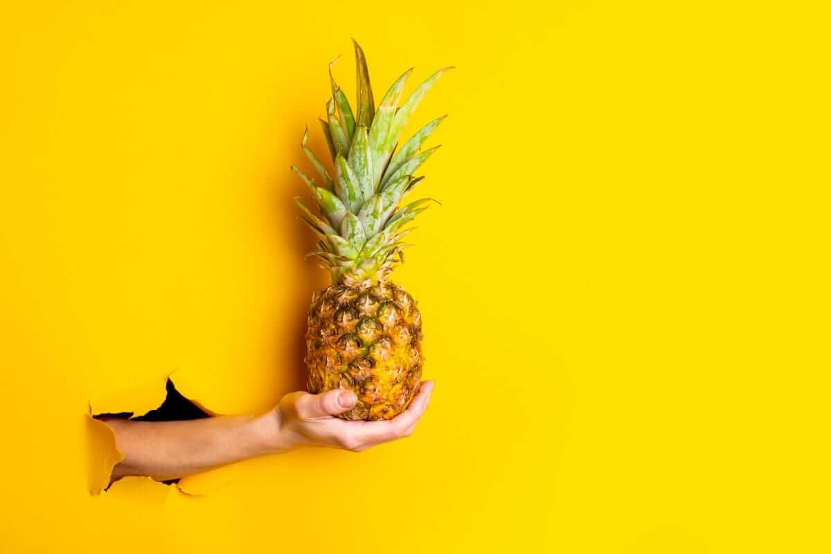 Ananas, Superfoods November - Mann hält Ananas in der Hand vor gelber Wand