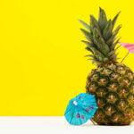 Ananas Mocktails (Frische Ananas mit bunten Cocktail-Schirmchen vor einem leuchtend gelben Hintergrund, symbolisierend tropische Mocktails und Sommerdrinks.)
