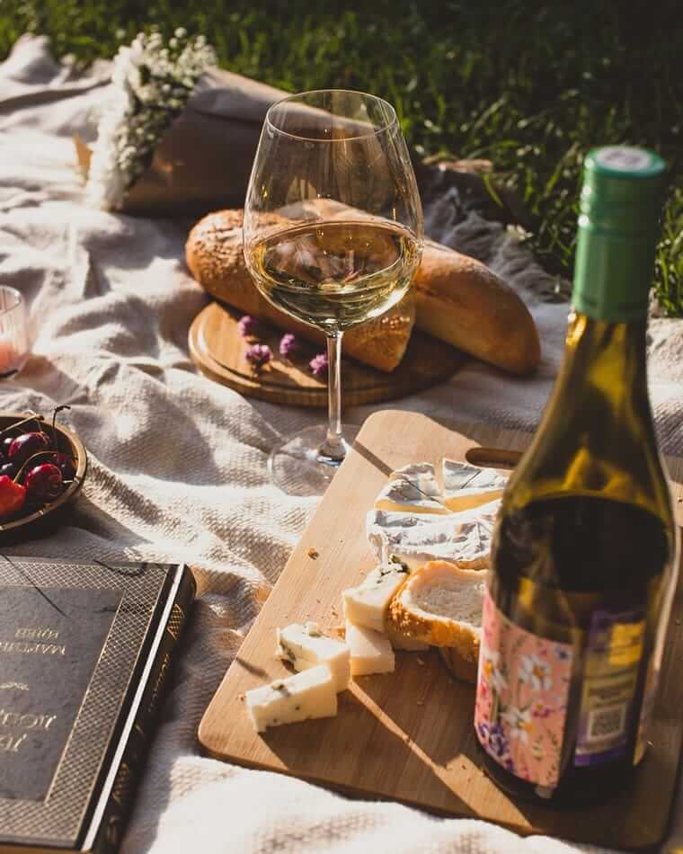 Käse und Wein (Picknick im Grünen mit einem Glas Weißwein, einer Flasche Wein, Käse, Baguette und Kirschen auf einem Holzbrett, umgeben von einer Decke, Blumen und einem Buch.)