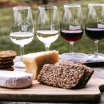 Käse und Wein (Holzbrett mit verschiedenen Käsesorten, knusprigem Saatenbrot und Crackern, dazu vier Weingläser mit Weiß- und Rotwein auf einem Holztisch im Freien.)