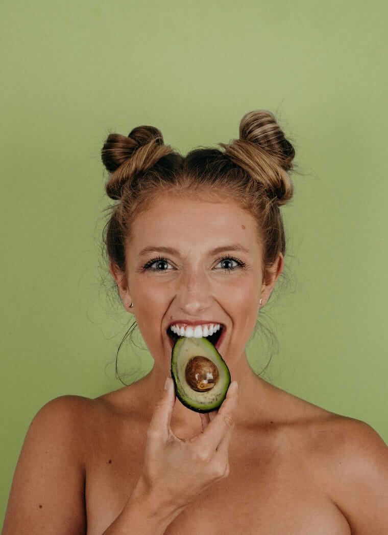 personalisierte Ernährung (Lachende junge Frau mit Dutt-Frisur hält eine halbe Avocado vor den Mund, grüner Hintergrund – Symbol für gesunde, individuelle und bewusste Ernährung.)