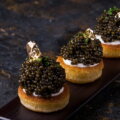 Kaviar / Drei Blinis mit Creme und schwarzem Kaviar, garniert mit essbarem Blattgold, serviert auf einem dunklen Teller vor schwarzem Hintergrund.