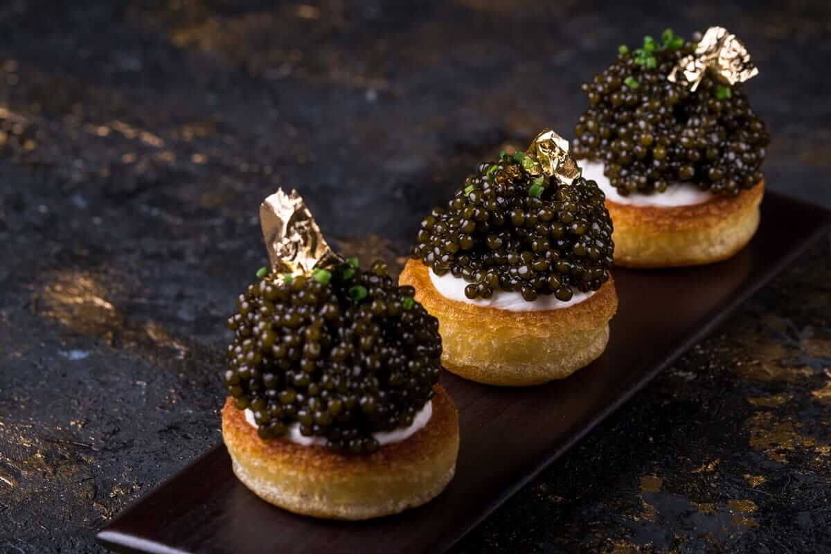 Kaviar / Drei Blinis mit Creme und schwarzem Kaviar, garniert mit essbarem Blattgold, serviert auf einem dunklen Teller vor schwarzem Hintergrund.