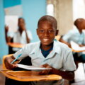 Essen mit Haltung / Ein afrikanischer Schüler im Klassenzimmer lächelt in die Kamera und hält ein Heft und einen Stift – Symbol für Bildung und Zukunftschancen durch faire Bedingungen.