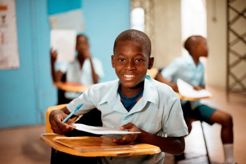 Essen mit Haltung / Ein afrikanischer Schüler im Klassenzimmer lächelt in die Kamera und hält ein Heft und einen Stift – Symbol für Bildung und Zukunftschancen durch faire Bedingungen.