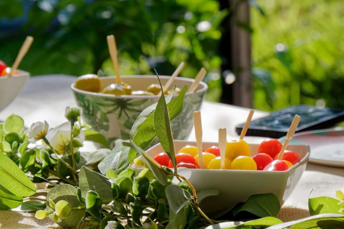 Sommer, Abnehmen (Sommerliche Snacks mit bunten Tomaten und Oliven in Schälchen auf einem hell gedeckten Tisch im Grünen – leichtes, gesundes Essen im Sonnenschein)