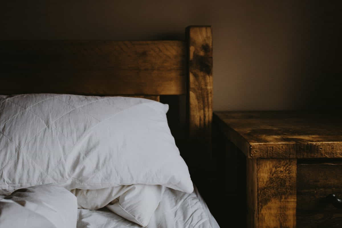 Bausteine für einen gesunden Schlaf: Vitamine, Mineralstoffe & mehr Schlaf / Holzbett mit weißem Kopfkissen und gedämpftem warmem Licht – ein ruhiger, gemütlicher Schlafplatz