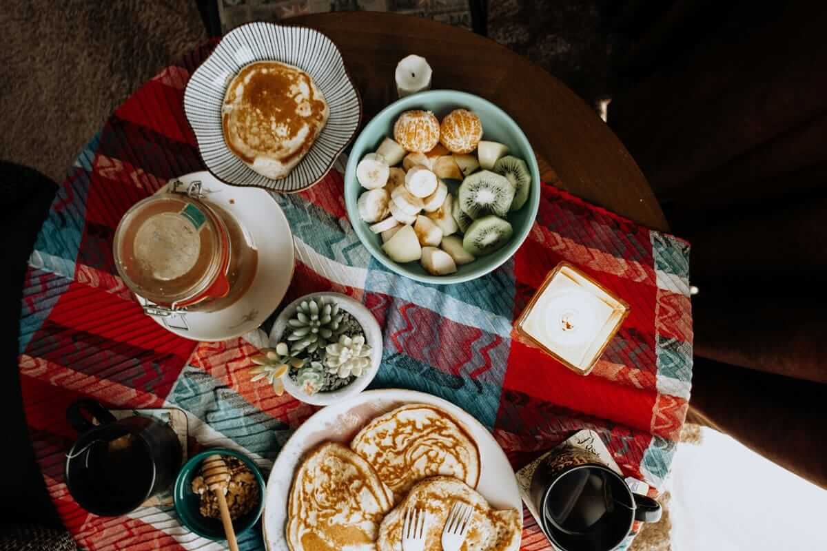 Gemütlich gedeckter Frühstückstisch mit Pancakes, frischem Obst, Honig und Kerze auf einer bunten Decke als warmes Mood-Food-Arrangement.