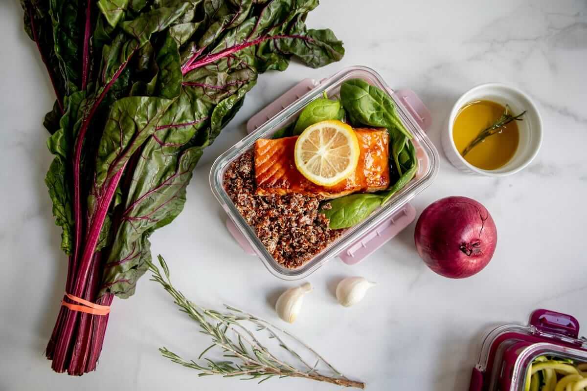 Warum Lieblingsgerichte nachhaltiger sind als neue Rezepte Lieblingsgerichte / Lunchbox mit Lachs, Quinoa und Gemüse neben frischem Mangold und Gewürzen – einfache Zubereitung im Alltag