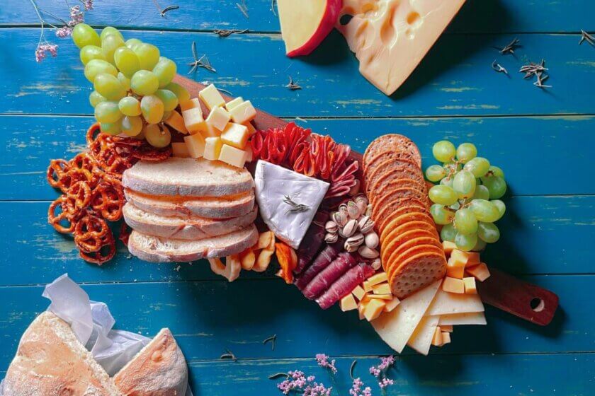Kombinieren statt verbieten / Verschiedene Lebensmittel wie Brot, Käse, Trauben, Nüsse, Aufstriche und Avocado auf einem Holzbrett als Beispiel für ausgewogenen Genuss durch Kombination