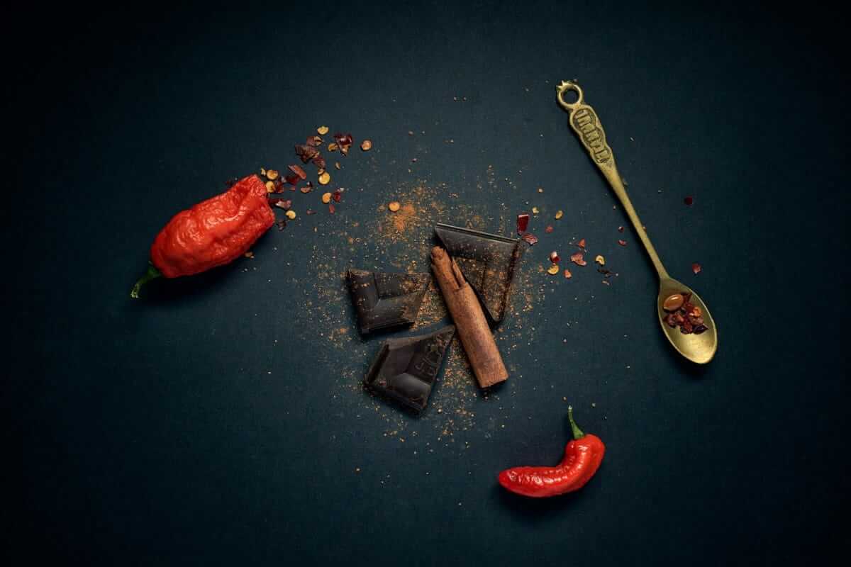 Swicy Food: Wenn süß auf scharf trifft – und warum das nicht verrückt ist Dunkle Schokolade, rote Chilischoten, Zimtstangen und Chilipulver auf dunklem Untergrund – Zutaten für Swicy Food