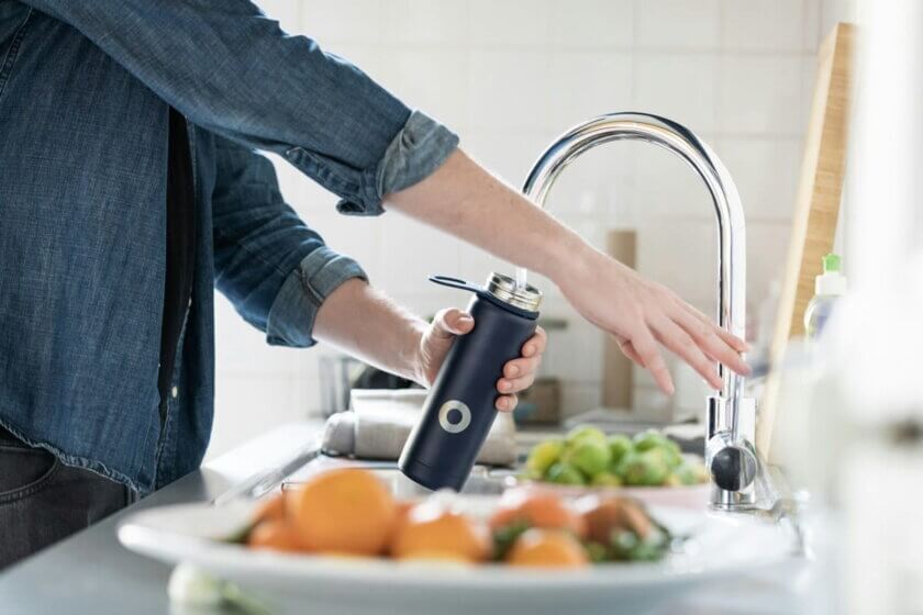 Leitungswasser / Person füllt wiederverwendbare Trinkflasche mit Leitungswasser am Küchenhahn – nachhaltige Alternative zu Mineralwasser