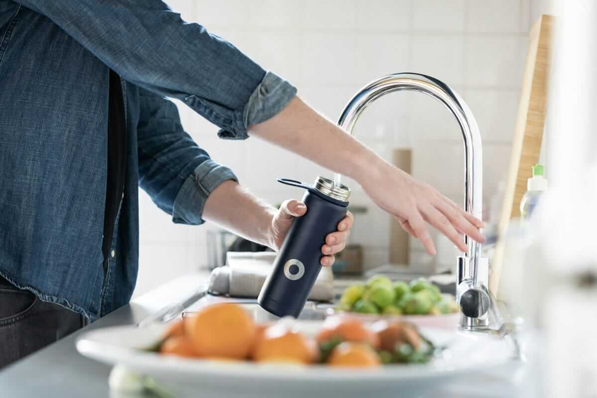 Warum Leitungswasser die unterschätzte Nachhaltigkeits-Entscheidung ist Leitungswasser / Person füllt wiederverwendbare Trinkflasche mit Leitungswasser am Küchenhahn – nachhaltige Alternative zu Mineralwasser