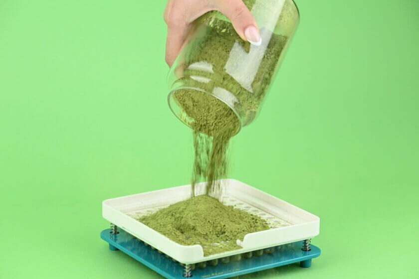 Füllstoffe / Hand schüttet grünes Pulver aus einem Glasgefäß auf eine Kapselabfüllform – Herstellung von Nahrungsergänzungsmitteln in Pulverform.