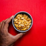 Snacken / Hand hält eine Schüssel mit gerösteten Kürbiskernen vor rotem Hintergrund
