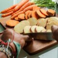 Hände schneiden Süßkartoffeln und Karotten in Scheiben auf Holzbrett – Vorbereitung für Meal-Prep