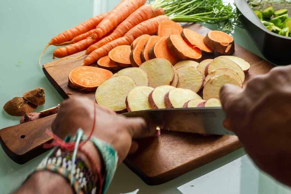 Einfaches Meal-Prep: Drei Techniken statt Großvorbereitung Hände schneiden Süßkartoffeln und Karotten in Scheiben auf Holzbrett – Vorbereitung für Meal-Prep