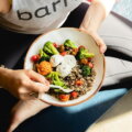 Lebensmittelkonsum / Draufsicht auf eine Bowl mit Quinoa, Brokkoli, Tomaten und Burrata in den Händen einer Person