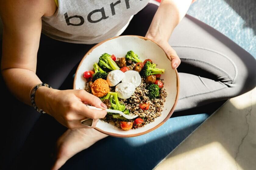 Lebensmittelkonsum / Draufsicht auf eine Bowl mit Quinoa, Brokkoli, Tomaten und Burrata in den Händen einer Person