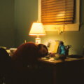 Erschöpfung / Person mit dem Kopf auf dem Schreibtisch, dahinter eine Schreibtischlampe und unaufgeräumter Tisch am Abend
