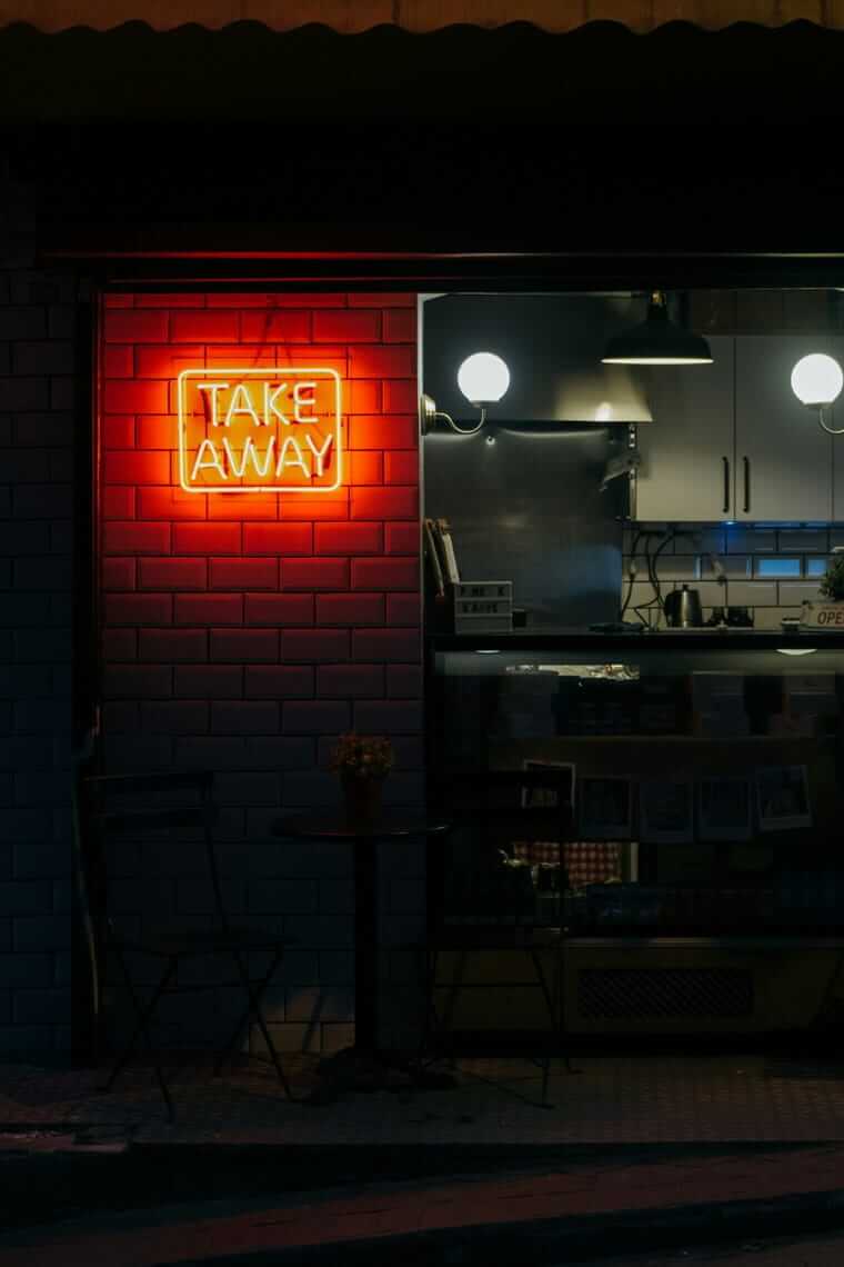 Wenn Überforderung auf den Teller schlägt: Warum bei Erschöpfung zuerst das Essen leidet Erschöpfung / Neonschild mit der Aufschrift „Take Away" an einer Backsteinwand bei Nacht