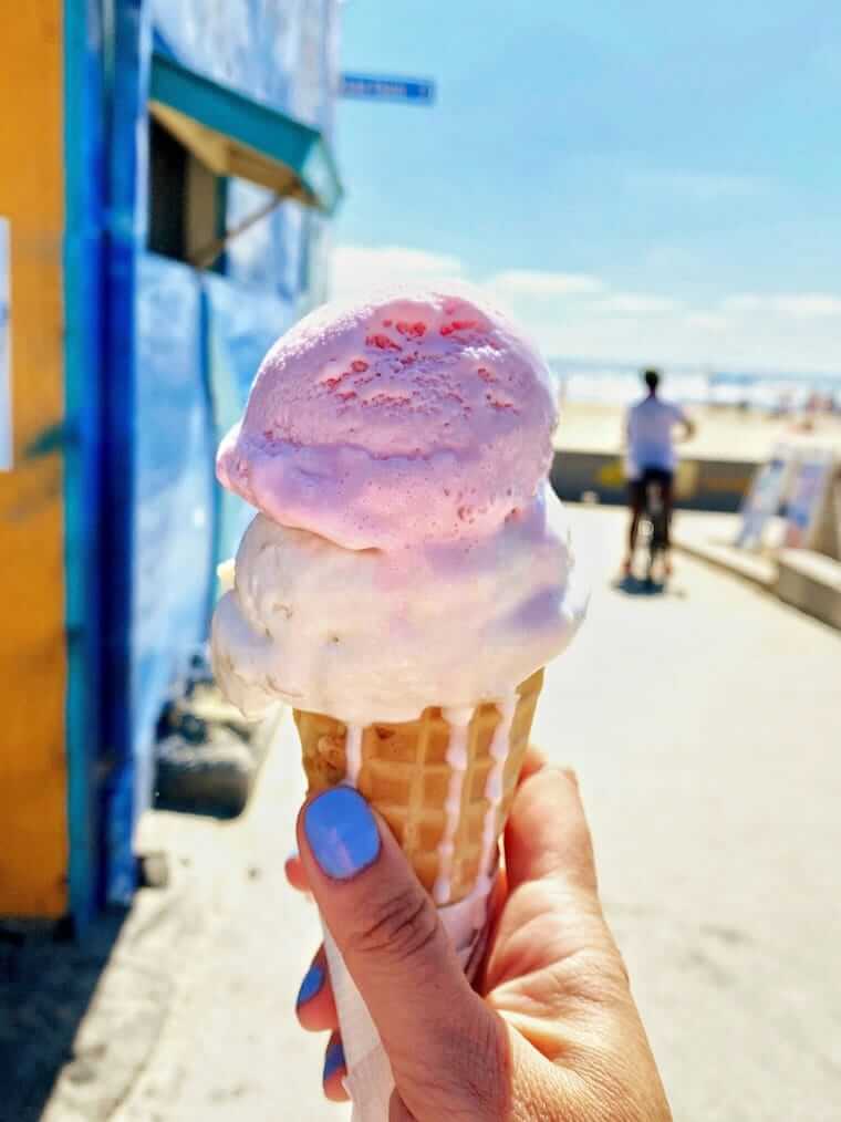 Hand hält eine schmelzende Eistüte mit rosa und weißer Kugel vor sommerlicher Straßenkulisse