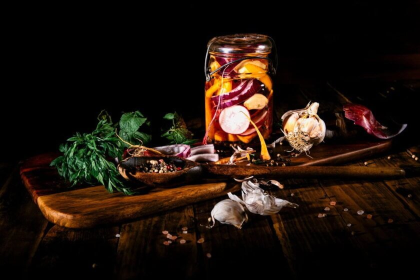 Eingelegtes Gemüse im Glas mit Knoblauch und Kräutern auf einem Holzbrett – Haltbarmachen als Weg gegen Lebensmittelverschwendung im Sommer.