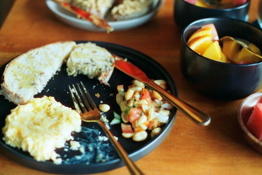 Kleines Frühstück auf schwarzem Teller – Rührei, Toast und eine Schüssel mit frischem Obst, aufgenommen bei warmem Tageslicht