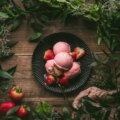 Rosa Eiskugeln mit frischen Erdbeeren auf einem dunklen Teller, umrahmt von grünen Zweigen