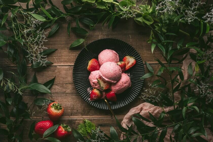 Rosa Eiskugeln mit frischen Erdbeeren auf einem dunklen Teller, umrahmt von grünen Zweigen