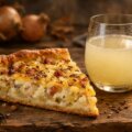 Ein Stück warmer Zwiebelkuchen mit Kümmel neben einem Glas Federweißer auf rustikalem Holztisch