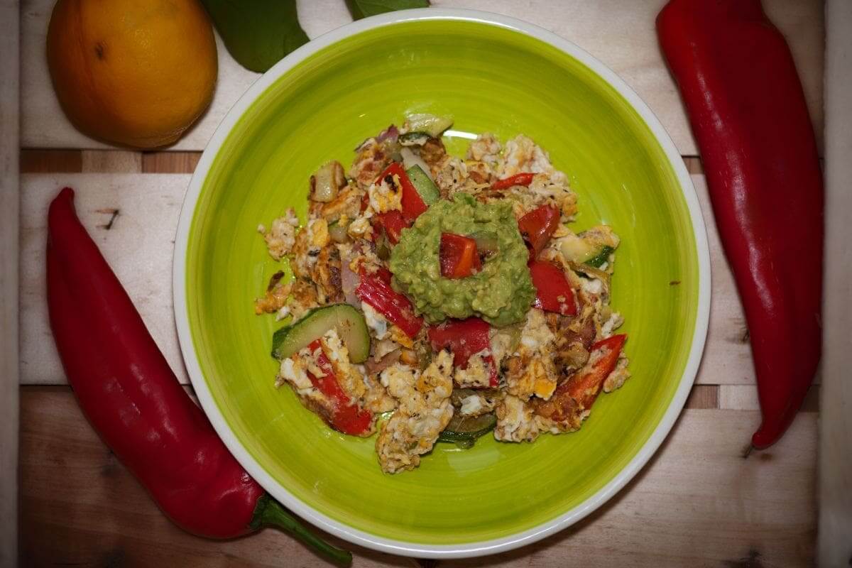 Rührei mit roten Paprikastücken und Guacamole in grüner Schüssel, daneben rote Spitzpaprika